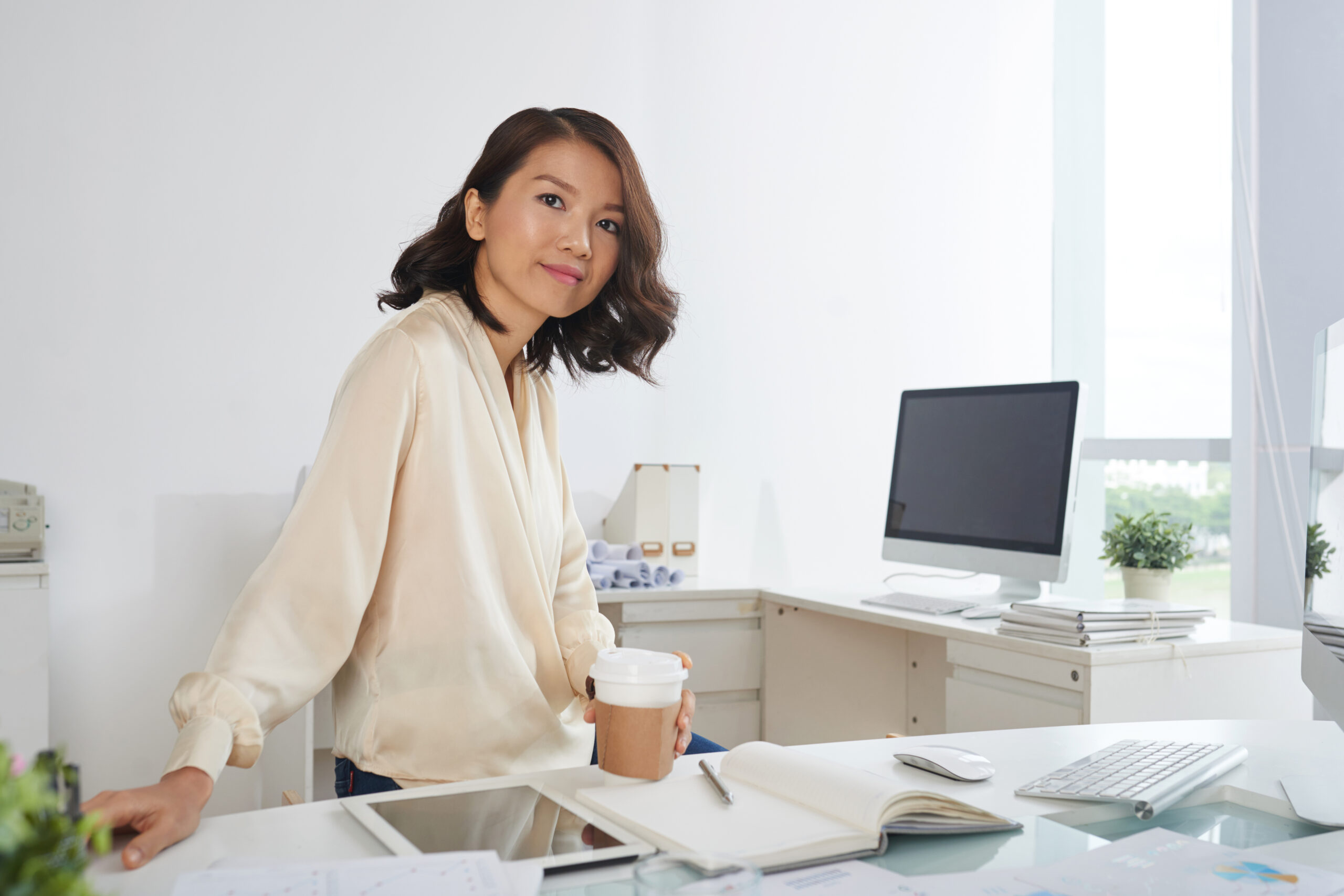 Portrait of young Asian businesswoman posing at workplace with paper coffee cup